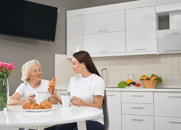 Front view of smiling relatives drinking tea and spending time together in white kitchen. Young brunette laughing and showing croissant to happy grandmother. Concept of family.